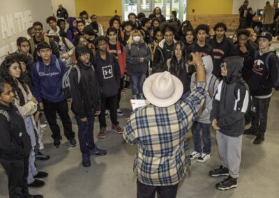A man in a checkered shirt and white hat greets a large group of students