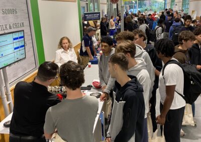 High School students gather around exhibitor tables in a long hallway.